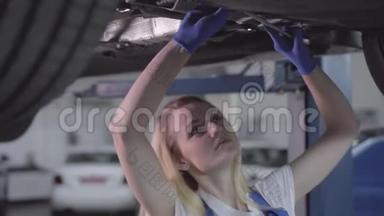年轻的白种人女人站在车下，拧紧螺丝。 在修理厂工作的金发女汽修工